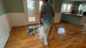Professional contractor using buffing machine for hardwood floor screening in Denver mountain home showing contrast between refinished glossy surface and dull unfinished area during screen and recoat process